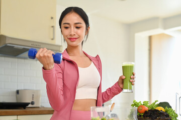 Attractive woman drinking fresh green detox vegetable juice and lifting dumbbell in kitchen. Healthy lifestyle concept