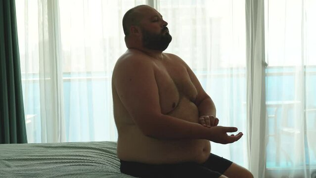 Overweight man measures pulse on hand, doing breathing exercises during seizure sitting in bed. Breath difficulties, asthma, postcovid complications. Health problems of excess weight people concept.