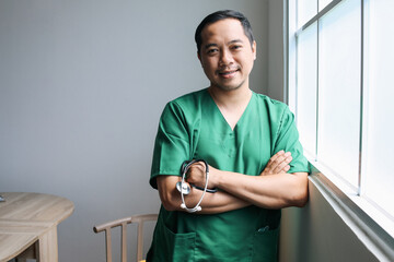 Portrait of smiling Asian male doctor posing with folded arms and standing near window at hospital. 