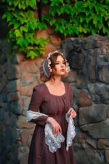 Beautiful red haired girl in brown long medieval dress and bonnet standing near stone wall. Art...