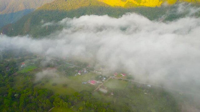 Morning in the village of Oxapampa, Peru, captured from a high-angle drone perspective. A 180-degree rotating shot reveals the village shrouded in clouds, with houses, lush fields, and hills in view.
