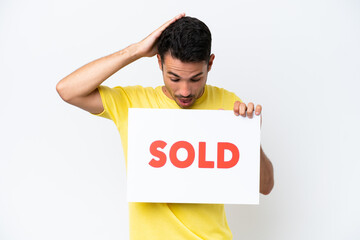 Young handsome man over isolated white background holding a placard with text SOLD with surprised...