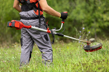Man mowing tall grass with electric cordless lawn trimmer in backyard. Garden care tools and...