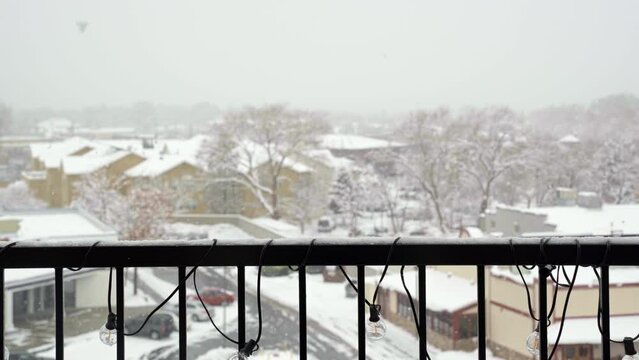 Medium shot of snowflakes falling during a old winter snowstorm with an apartment balcony railing and other buildings, trees, and a parking lot out of focus in the background in Midvale Utah