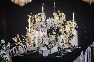 Luxury wedding reception. Trendy decor large chandelier for a birthday party. Table setting, setup. Banquet decoration composition flowers, candles in hall restaurant. Table covered black tablecloth.