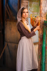 Fototapeta premium Beautiful young rural girl in medieval dress and bonnet hat posing on a ranch. Pretty blonde model in countryside art photo. 