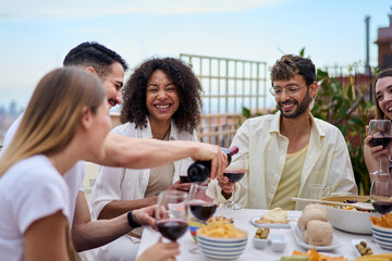 Young smiling Caucasian man serving red wine to guests at food table in celebration with happy friends on rooftop. Group of cheerful multiracial friends gathered for lunch party on outdoor terrace.