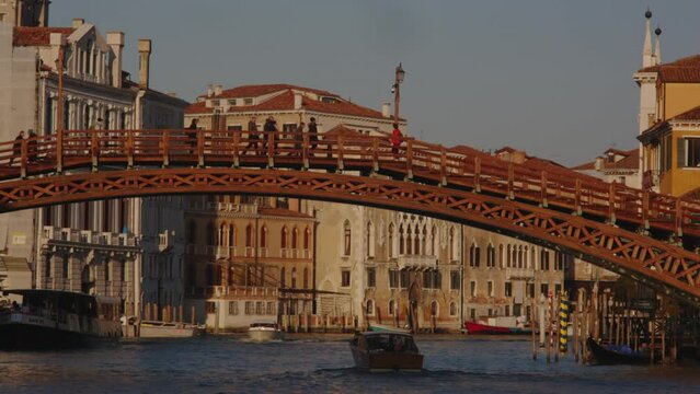 Boats passing underneath the Accademia Bridge in Venice as tourists walk over with the Grande Canale palatial buildings in the background