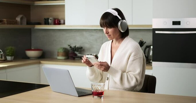 A Caucasian woman, wearing headphones, efficiently juggles texting on phone and working with laptop in the kitchen. Her ability to balance communication with work tasks creates a dynamic atmosphere.