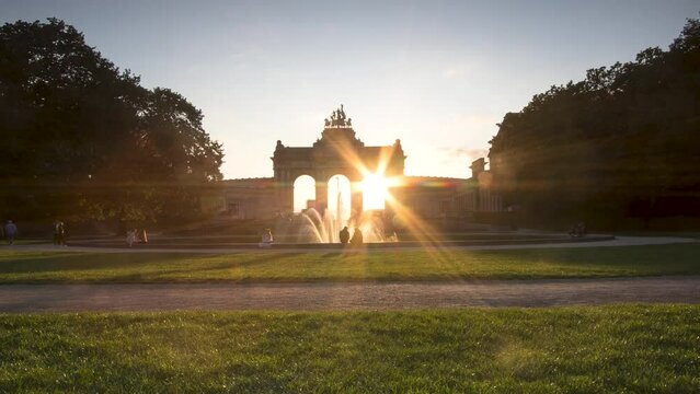 Romantic sunset time lapse of the Cinquantenaire Park with the fountain and the lights turning on as the sun goes down. Lovers and visitors having a picnic in the large park with clouds and many color