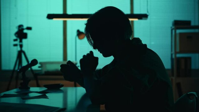 Medium silhouette video of a suspect, offender, perpetrator or prisoner sitting in the interrogation room. He is studying the evidence on the table.