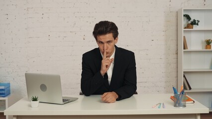 Medium shot of a young man sitting at the table in the office, thumbing his lips, shushing as if he just told you a secret. He looks sneaky and serious.