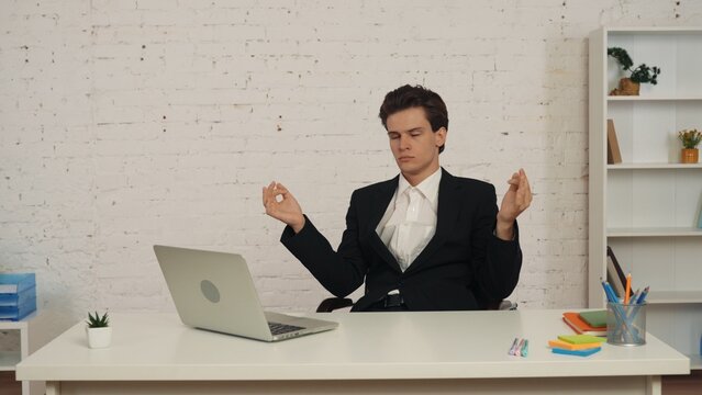 Medium shot of a young man sitting at the table, holding hands up in metitation gesture as if he is trying to calm down. He looks peaceful with his eyes closed.