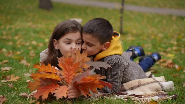 Charming boy and girl hugging in slow motion kissing cheeks lying on autumn lawn. Portrait of carefree happy Caucasian friends enjoying leisure in park on overcast day