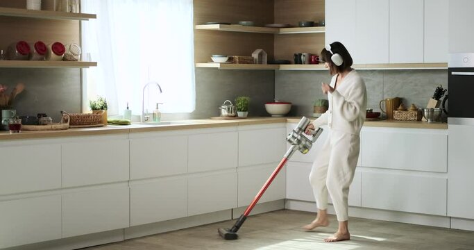 A carefree Caucasian woman, wearing headphones, combines household chores with entertainment as she vacuums and dances in the kitchen. Her joyful and uninhibited movements create a fun atmosphere.