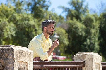 A handsome young man with a beard and sunglasses in his hand is leaning on the railing of a park....