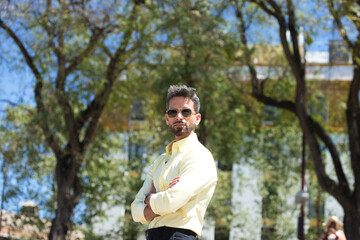 Naklejka na ściany i meble handsome young man with beard and sunglasses, arms folded and confident. The man is in a garden in seville with trees in the background.