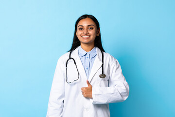 Fototapeta premium Smiling Indian Doctor Young Woman Gesturing Thumbs Up, Blue Background