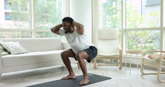 Strong African muscular man wear sportive t-shirt and shorts practicing squats standing on mat training alone in cozy living room. Healthy lifestyle, exercises for good shape, home work out activity