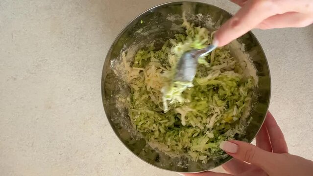 The girl is using a spoon to mix together grated zucchini, eggs, and flour in an iron bowl, to prepare zucchini fritters or zucchinipizza. zucchini recipes