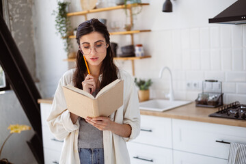 Woman in eyeglasses reading book and holding pencil in kitchen at home, self-improvement concept 