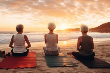 Poster A group of senior happy woman is doing yoga exercises relaxed and mindfull with a yoga mat on a beautiful beach at sunset © pangamedia