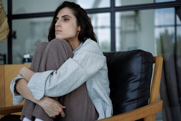 Disappointed brunette woman hugging knees and looking away while sitting on armchair at home