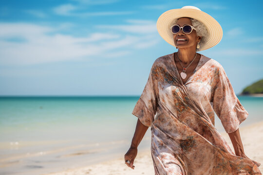 A senior old beautiful african american woman is walking on the sand next to the waterline with a dress on a tropical beach with a calm ocean - spring weather beach relaxing - Powered by Adobe