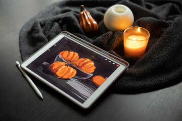 The still life picture with pumpkins drawn on the electronic tablet next to burning candle and ceramic pumpkin figurine on black wooden table. The concept of inspiration, creativity, self-development,