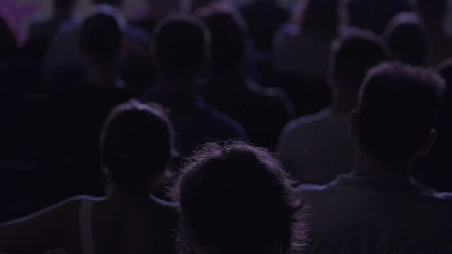 Tilt up view of crowd sitting on seats against stage in dark hall during conference and listening to performance attentively
