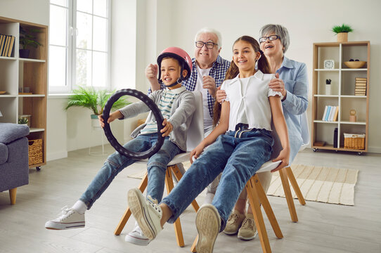 Happy grandparents and children play games together. Funny grandfather, grandmother, little girl, and boy in helmet with steering wheel sit on chairs, imagine to be in car and pretend to drive away
