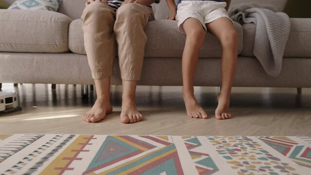 Wireless robot vacuum cleaner removes dust from parquet while mother and child rests on sofa in living room closeup. Automatic appliance for chores. Raise their legs to allow robot to clean