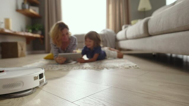 Wireless robot vacuum cleaner removes dust from parquet while mother and child rests on floor on carpet in living room. Automatic appliance for chores. Technology makes life easier. 