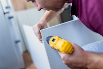 Man holding electrical screwdriver while assembling kitchen wooden elements.