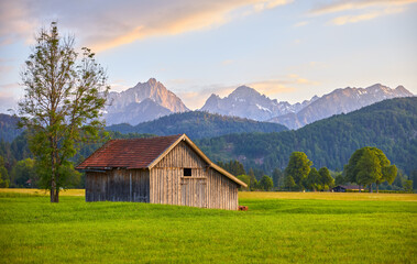 Fototapeta na wymiar Wooden barn on meadow in Bavaria, Germany. Panorama of green field with grass and picturesque german Alps mountains during sunset