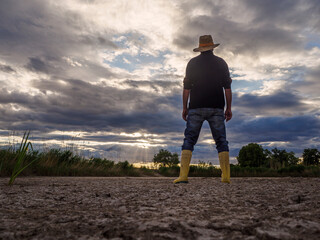 With rubber boots for the dry field. The farmer goes with his rubber boots and his shovel to a barren field. View from behind. Drought concept.