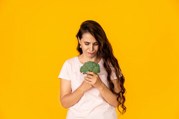 Sad young woman rejecting healthy broccoli on yellow color background. Diet concept
