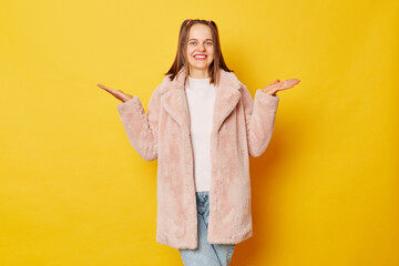 Glad optimistic brown haired girl wearing pink fur coat with ponytails raises both palms, pretends holding two items, being in good mood isolated over yellow background holding copy space