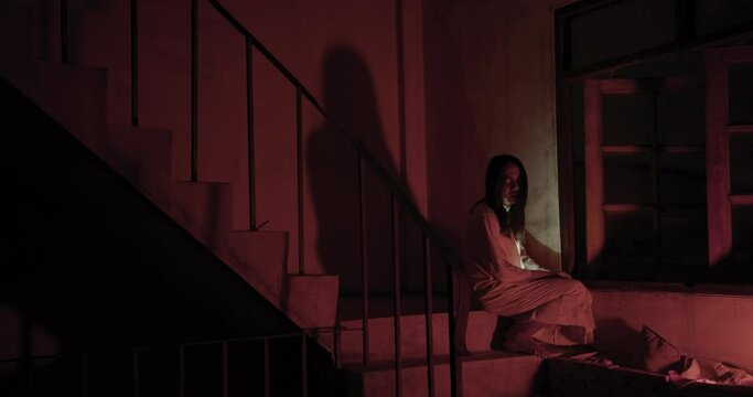 Horror scene of a mysterious Scary Asian ghost woman creepy have hair covering the face sitting on staircase at abandoned house with background dark red scene movie at night, holiday ghost Halloween