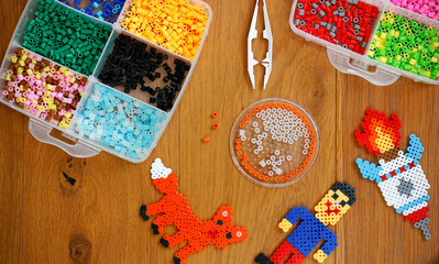 Colorful iron beads (perler or fused beads) in a box, art toys created from them and tweezers on wooden background. Toy developing children's imagination. Concept of children's creativity. Top view