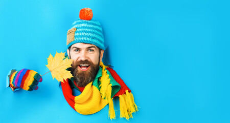 Smiling bearded man in colorful knitted hat, scarf and gloves looking through paper hole. Handsome guy in warm wear with yellow maple leaf. Winter time. Season sale. Discount. Copy space. Advertising.