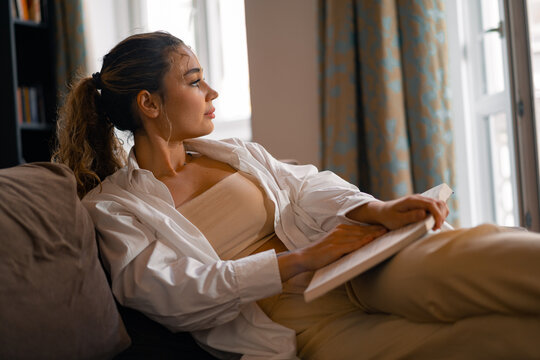 Young attractive woman looking at window while reading interesting book.