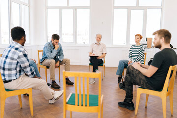 Wide shot portrait of senior male speaking on therapy meeting of addicts, sharing problem, giving support to teammates. Businessman, boss talking to colleagues, sharing ideas to project staff
