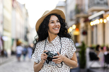 Young beautiful hispanic woman with curly hair walking in the evening city with a camera, female...