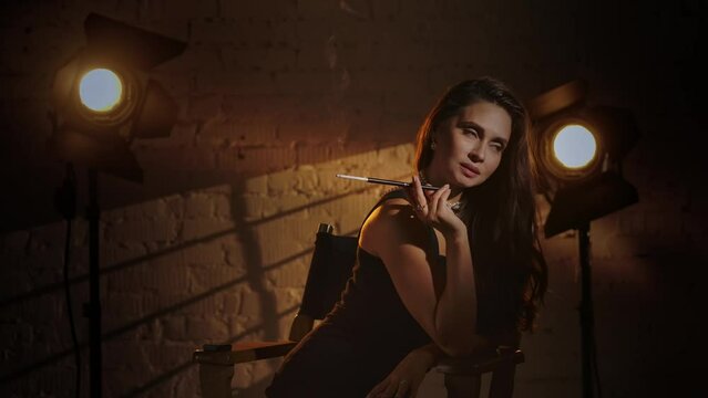 Elegant woman in black dress sitting on a director's chair, holding cigarette holder and smoking, smiling playfully looking at the camera.