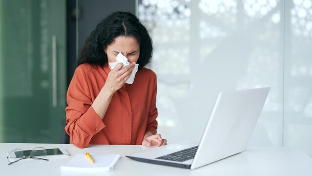 Young female employee with an allergy sneezes while sitting at a workplace at in modern office. Sickbrunette woman wipes her nose with a handkerchief. She has the first signs of a cold, virus, or flu