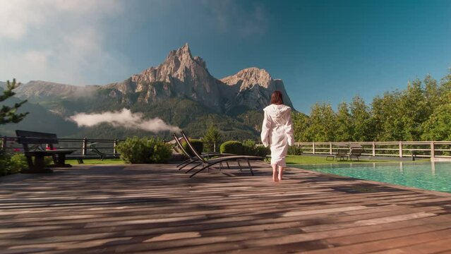 Woman in white robe walking to the infinity pool, enjoying luxury lifestyle on summer vacation at hotel spa with mountain view 