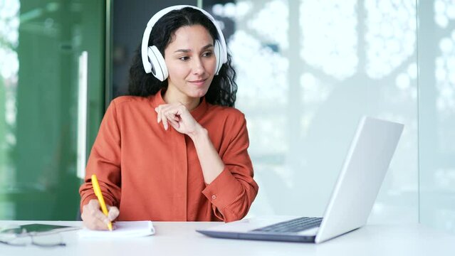 Young female employee in headphones watching video call conference notes in notebook looking at laptop sitting in office. Woman is studying online, listening remotely to a training course for workers