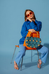 Fashionable young woman wearing trendy orange sunglasses, blue linen shirt, trousers, block heel shoes, holding straw wicker bag, posing on blue background. Studio portrait. Copy, empty space for text