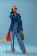 Fashionable young woman wearing trendy orange sunglasses, blue linen shirt, trousers, block heel shoes, holding bag, posing on blue background. Full-lenght studio portrait. Copy, empty space for text - 650386148